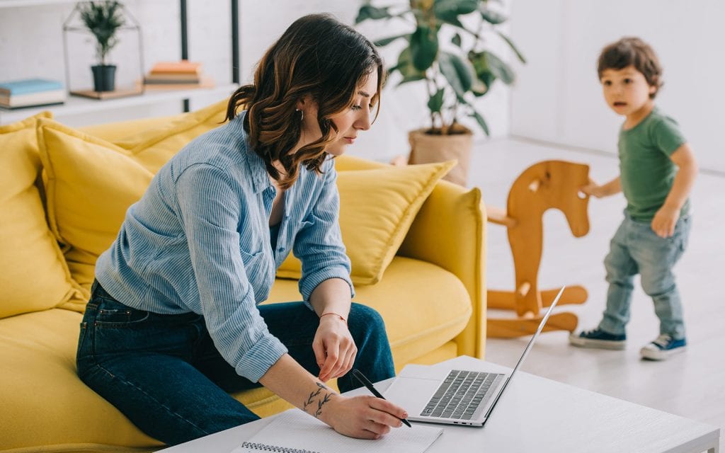 Woman working on laptop at home with toddler in the background